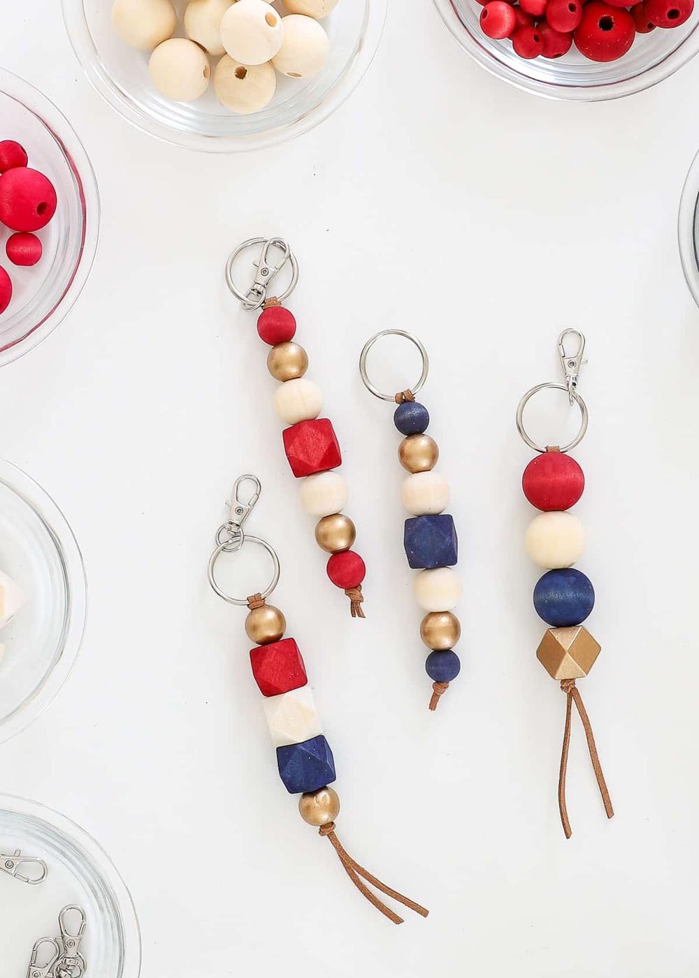 Three wood bead keychains in red, navy, cream and gold on key rings with suede tails, beside bowls of loose beads