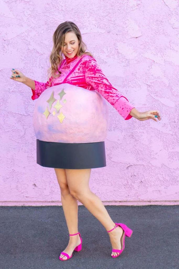 Adult wearing a pink crystal ball costume with gold stars and black base over a pink velvet top near a pink wall