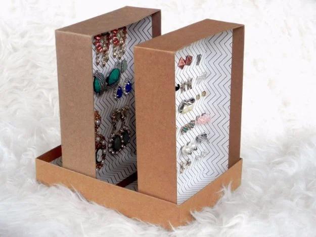 Two upright cardboard panels lined with hooked earrings, standing in a shallow box on a white fur surface