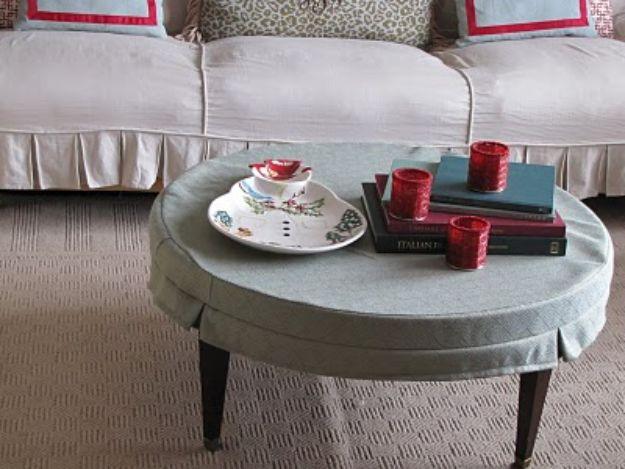 Oval coffee table under a grey green fabric slipcover, holding a tray of red cups and red books by a white sofa