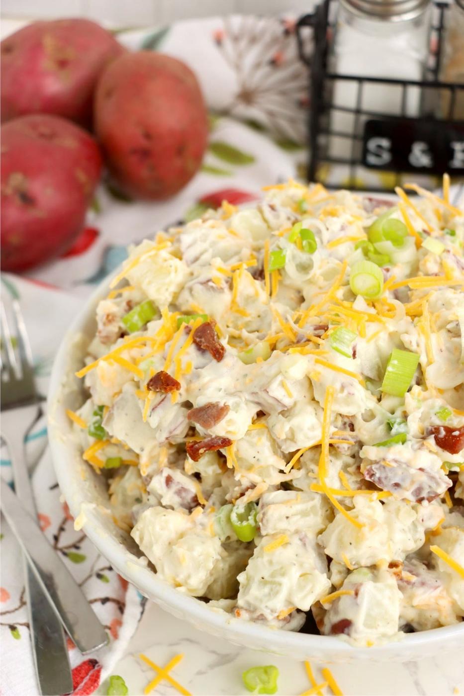 Loaded ranch potato salad in a white bowl with cheddar, bacon and green onion, red potatoes behind it