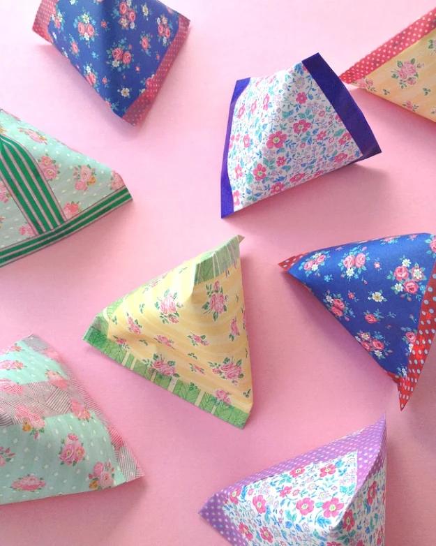 Pyramid gift pouches folded from floral and polka dot leftover wrapping paper, scattered on a pink background