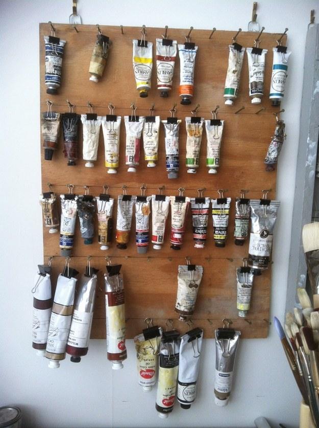 Rows of oil paint tubes hanging from binder clips on a wooden board in an artist's studio, labels facing out