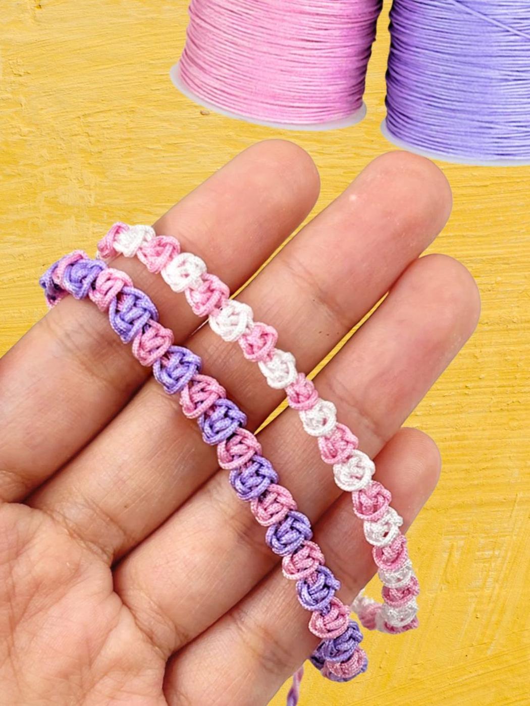 Pink, purple and white macrame rose pattern bracelet held over an open hand beside spools of colored cord