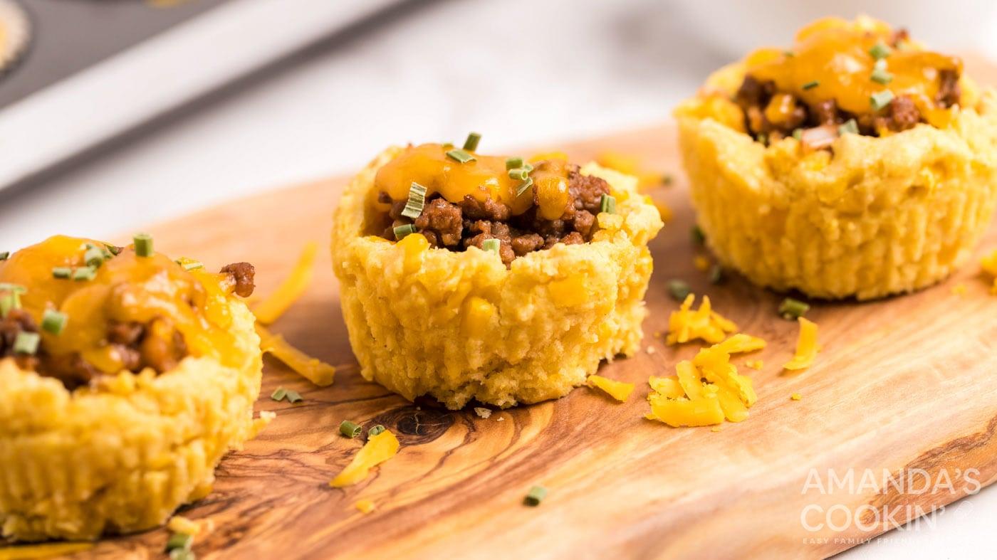 Sloppy joe cornbread muffins topped with melted cheddar and chives on a wooden board with shredded cheese