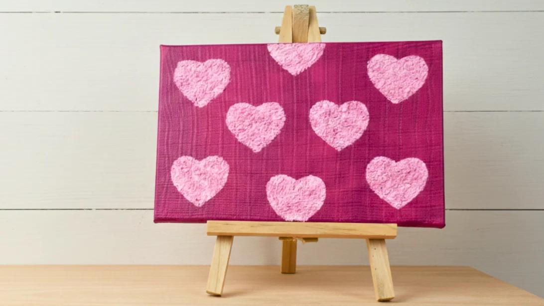 Small magenta canvas on a wooden easel with nine raised pale pink textured hearts made from tissue paper and paint