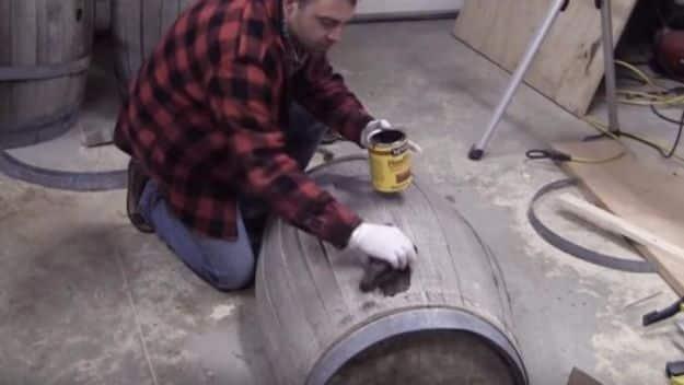 Whiskey barrel coffee table build, a man in a plaid shirt kneeling on a garage floor staining a barrel on its side