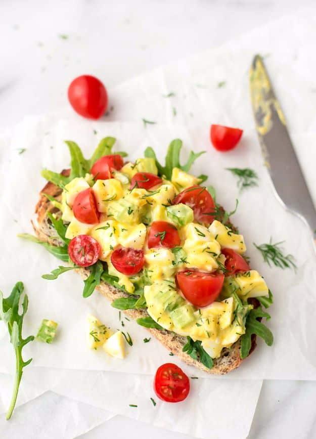 Healthy egg salad piled on one open faced toast with arugula, quartered cherry tomatoes, fresh dill and a knife