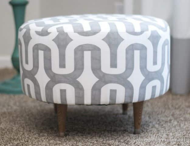 Round gray and white patterned upholstered spool ottoman on short wooden legs standing on beige carpet