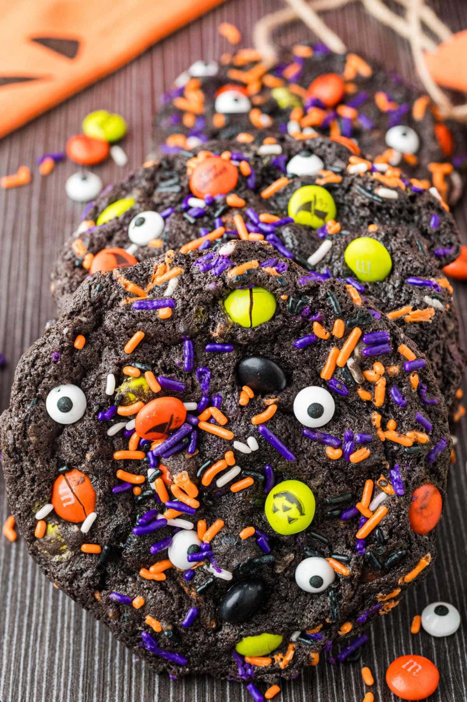 Chocolate Halloween cookies with candy eyes plus orange, purple and green candy pieces on a dark serving tray