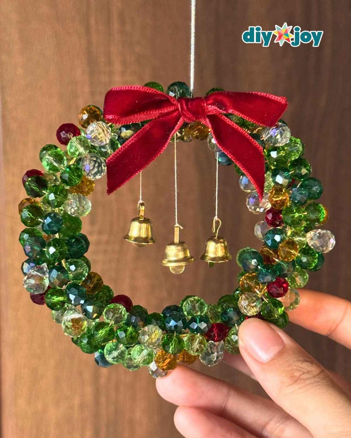 Beaded Christmas wreath ornament hanging against wood with a red velvet bow and three dangling gold bells