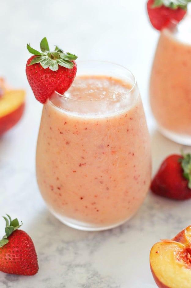 Pale pink strawberry peach smoothie in a stemless glass with a strawberry on the rim beside a fresh peach on marble