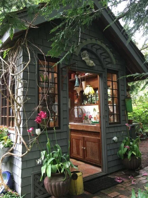 Rustic garden shed, a dark gray shingled shed with its doors open showing a wooden sink unit, plants and a stained glass lamp
