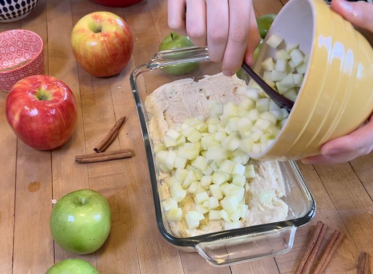 How to Make Apple Bread - hands pouring diced apples from a yellow bowl over the first batter layer in the glass loaf pan