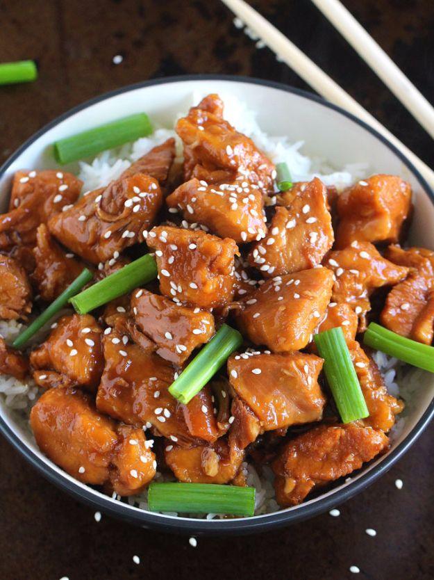 Dark bowl of glossy Mongolian chicken cubes over white rice with green onions and sesame seeds and chopsticks alongside