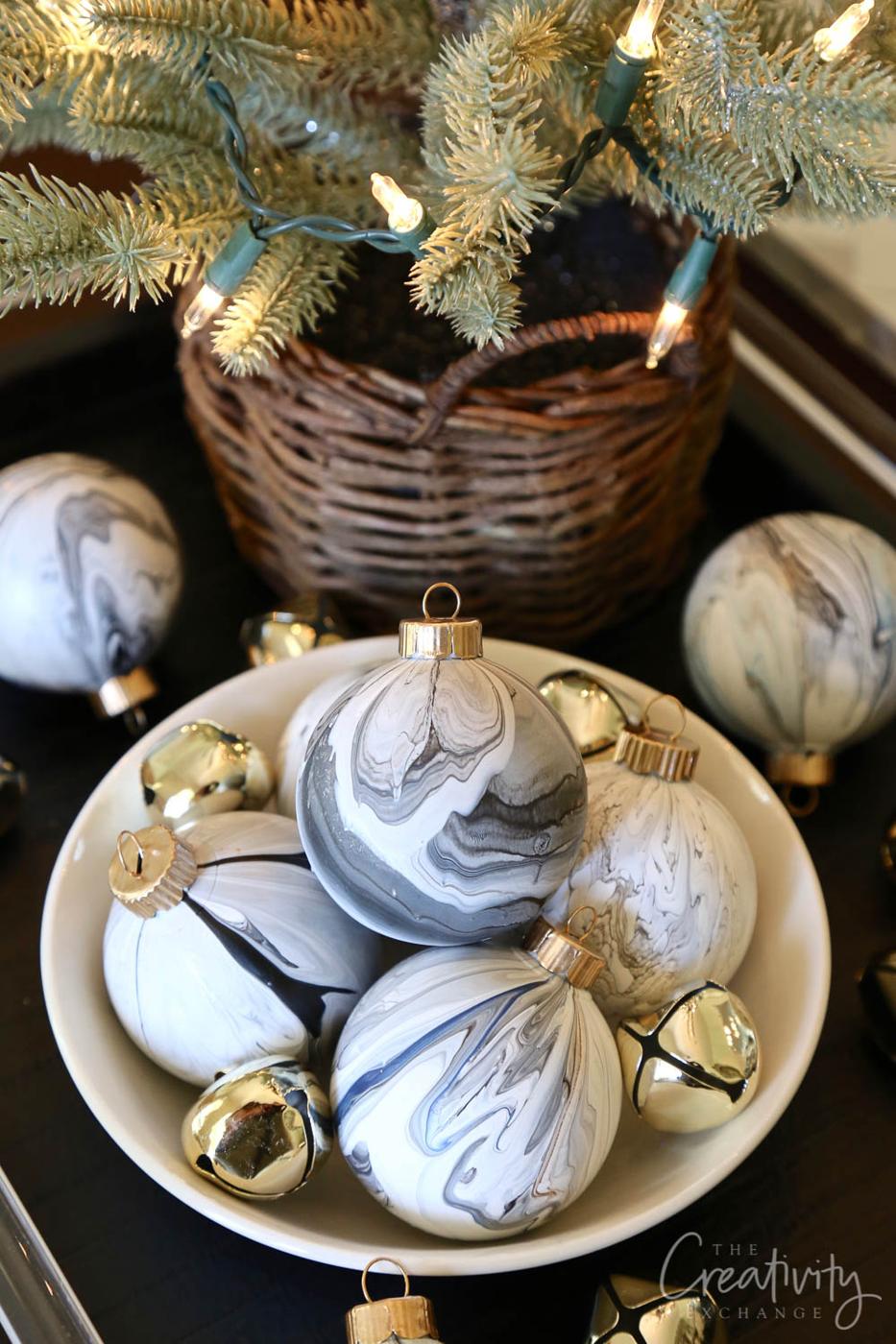 Gray and white marbled ball ornaments with gold caps piled in a white bowl with gold jingle bells under a lit mini tree
