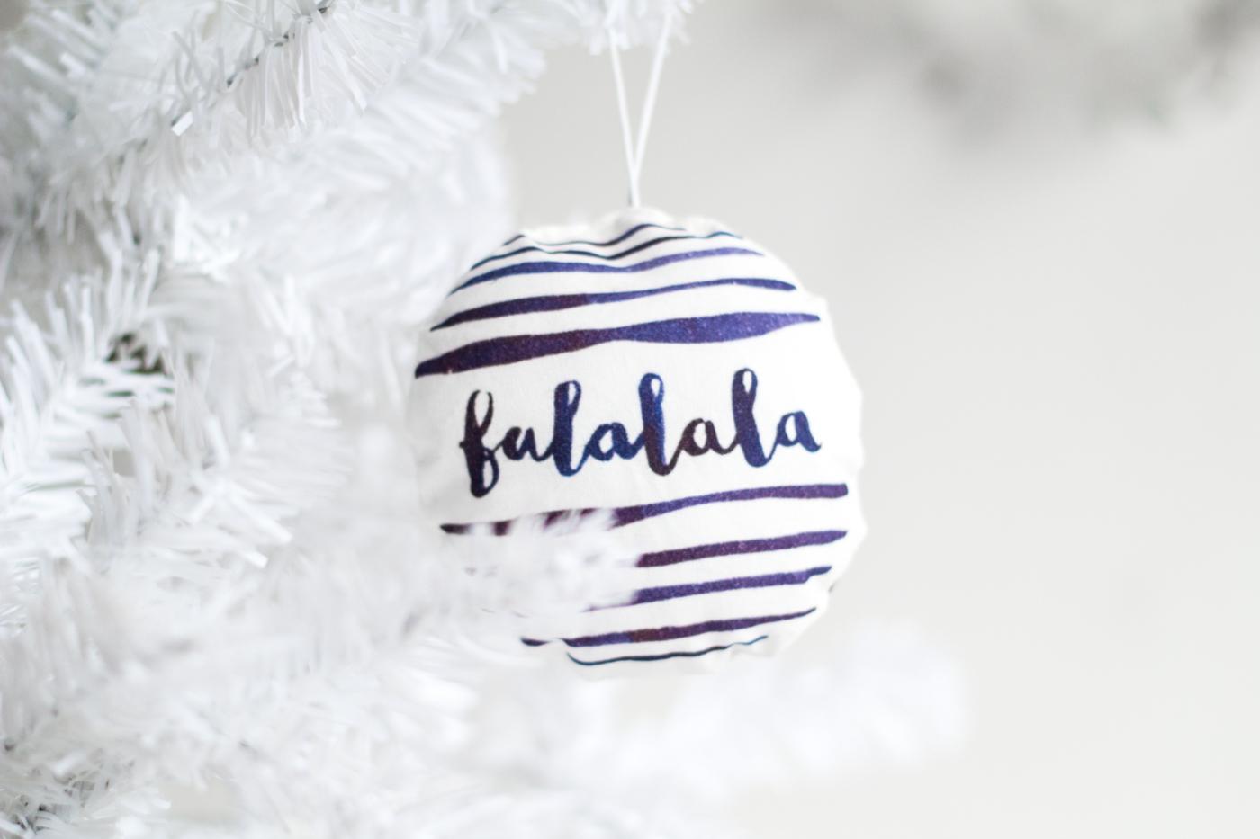 A soft stuffed fabric bauble printed with navy watercolor stripes and the words falalala hanging in a white Christmas tree