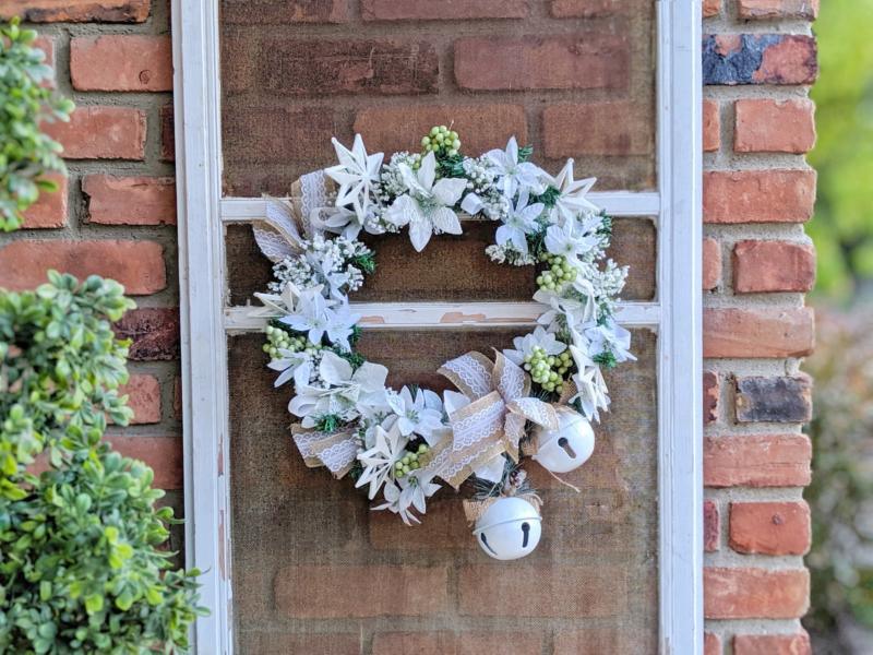 A wreath of white poinsettias, white stars, green berries, burlap and lace bows and two white bells on a window frame
