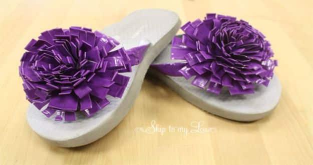 Pair of gray flip flops decorated with large ruffled purple duct tape flowers, photographed on a wooden floor