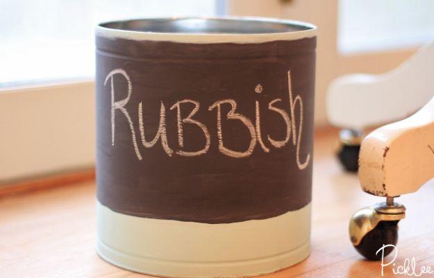 Popcorn tin trash can painted dark brown over cream with the word Rubbish in chalk, on a wood floor by a chair