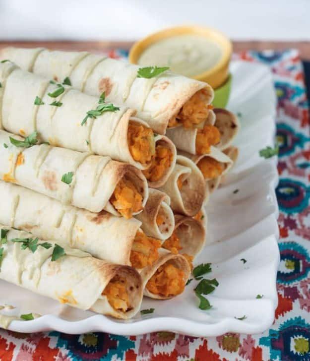 Baked buffalo chickpea and artichoke vegan taquitos stacked on a white platter with a dipping sauce bowl