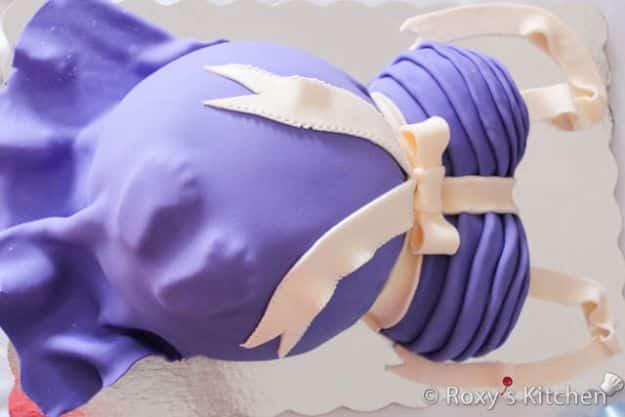 Baby shower cake shaped like a pregnant belly covered in purple fondant with a cream fondant ribbon and bow