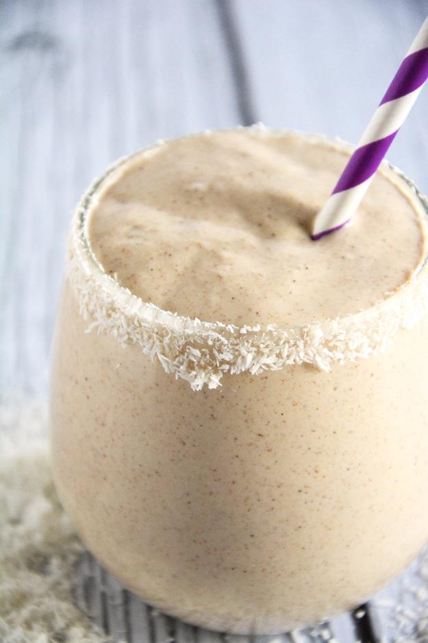 Creamy beige almond butter smoothie in a sugar rimmed glass with a purple striped straw on pale wood