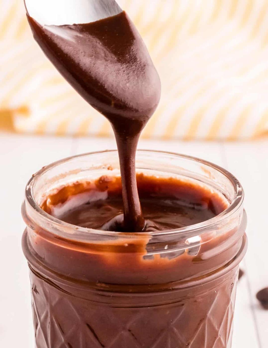 Glass jar of thick homemade hot fudge sauce with a spoon lifting a glossy chocolate drizzle on a white counter