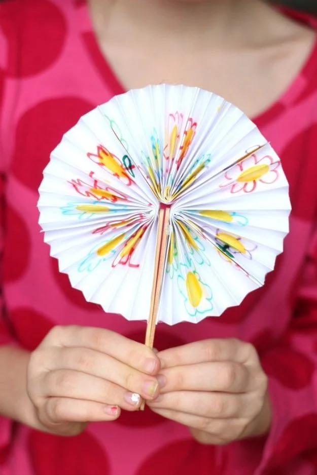 Child in a pink polka dot dress holding an open paper fan decorated with colored doodles, face not in frame