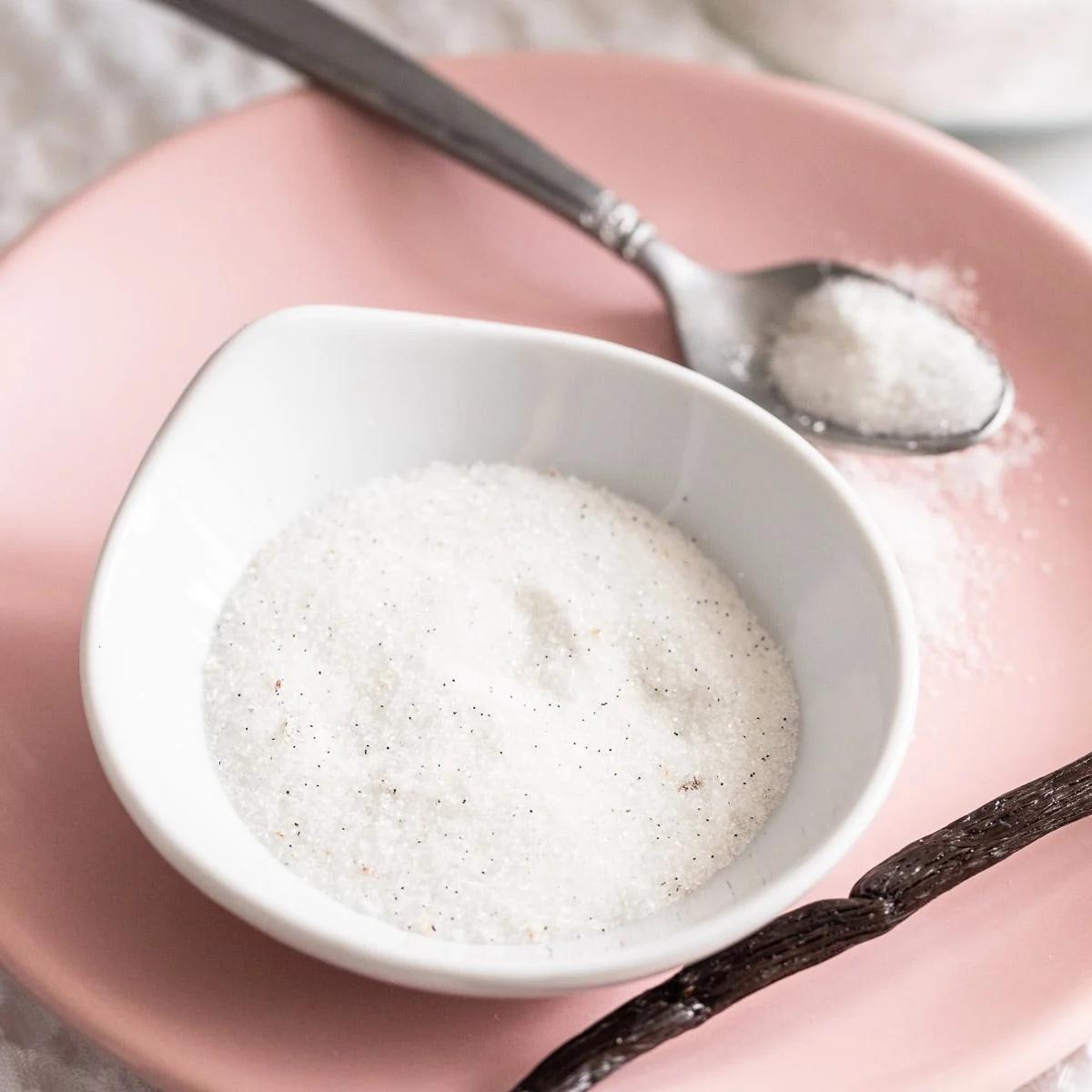 Small pink dish of white vanilla sugar with a silver spoon and a split vanilla bean resting on a pink plate
