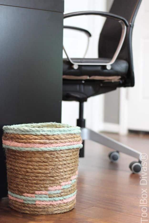 Rope wrapped bucket trash can painted with pink and mint bands, standing on a wood floor beside a black office chair
