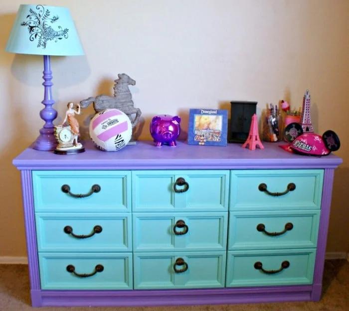 Kids dresser with mint green drawer fronts, a purple painted frame and black pulls, topped with toys and a lamp