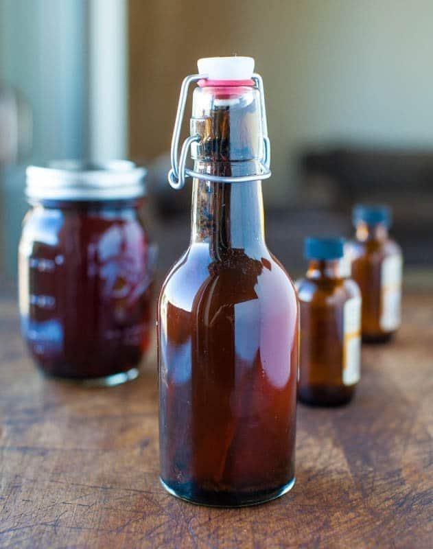 A dark amber swing top bottle of homemade vanilla extract on a wood table with a mason jar of beans behind it
