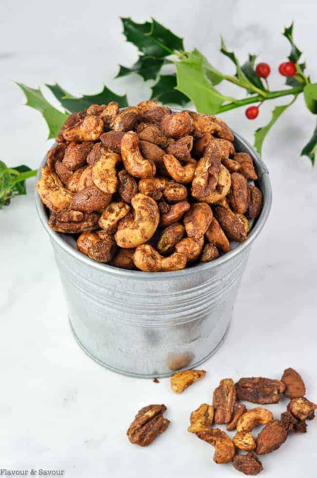 A small galvanized metal bucket heaped with gingerbread spiced mixed nuts beside a sprig of holly on white marble