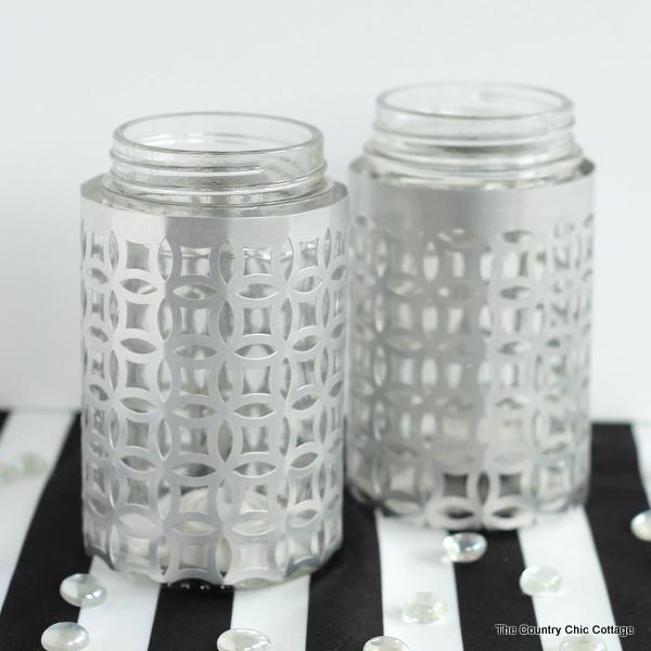 Two glass jars wrapped in silver lattice metal sleeves standing on a black and white striped surface