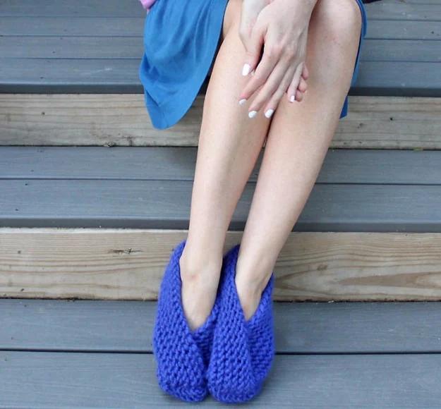 Bright blue knitted fold over slippers worn on bare feet on grey wooden decking, hands resting on the knees