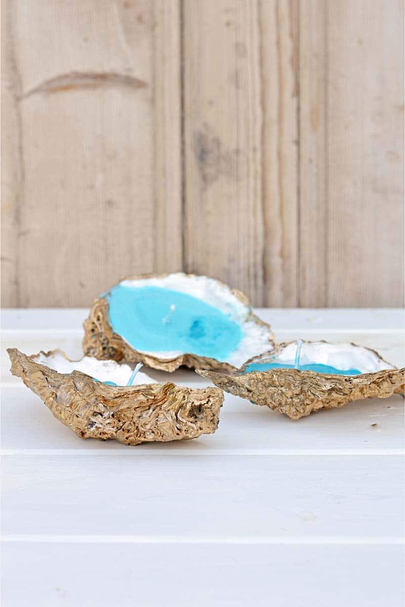 Gold painted oyster shells filled with turquoise wax and wicks, one propped on the other, on a white board