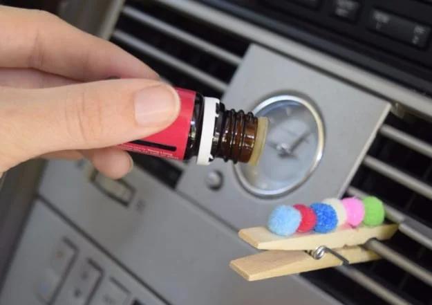 Wooden clothespin with pink, blue and green pom poms glued to it, clipped to a car dashboard air vent by a hand