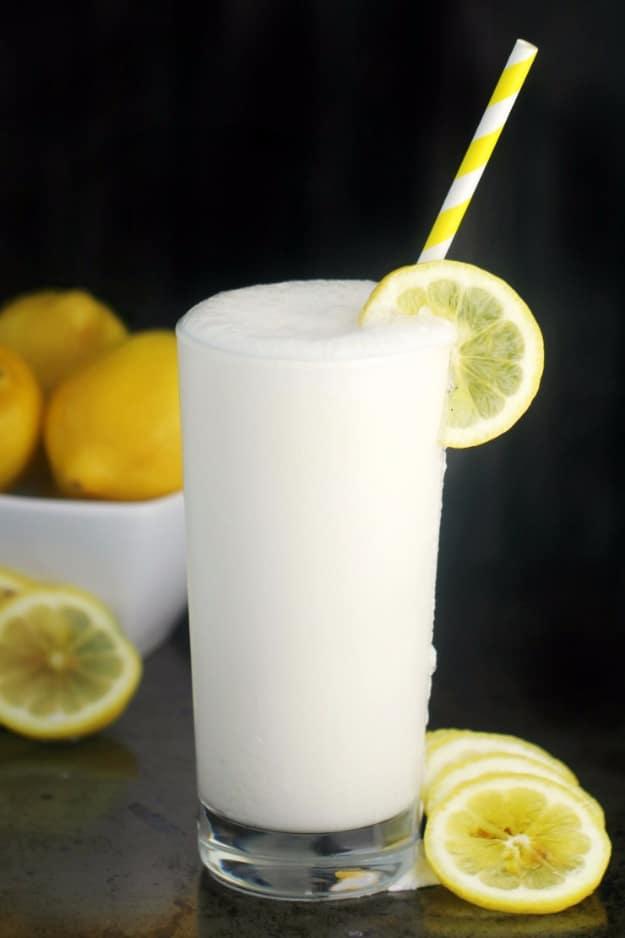 Tall glass of creamy pale frosted lemonade with a lemon slice on the rim and a yellow straw, lemons behind