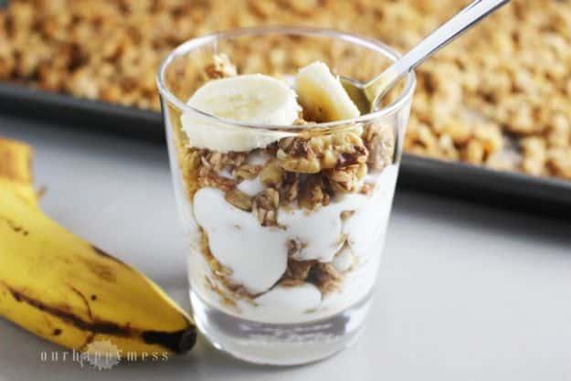 Glass of banana granola parfait with sliced banana beside a baking sheet of granola and whole bananas