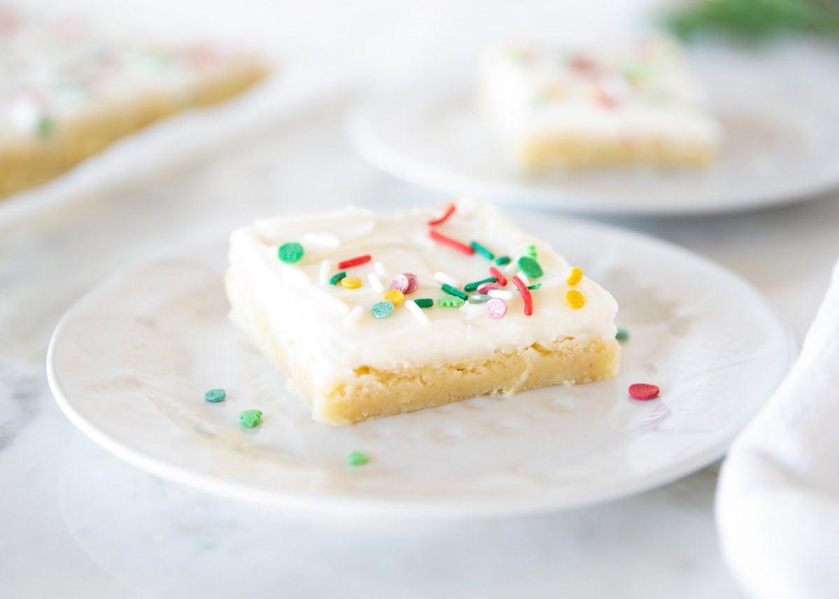 A square sugar cookie bar with thick white frosting and red, green and gold holiday sprinkles on a white plate