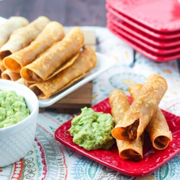 Oven fried chicken taquitos on white and red plates with one white bowl of guacamole and a stack of red plates