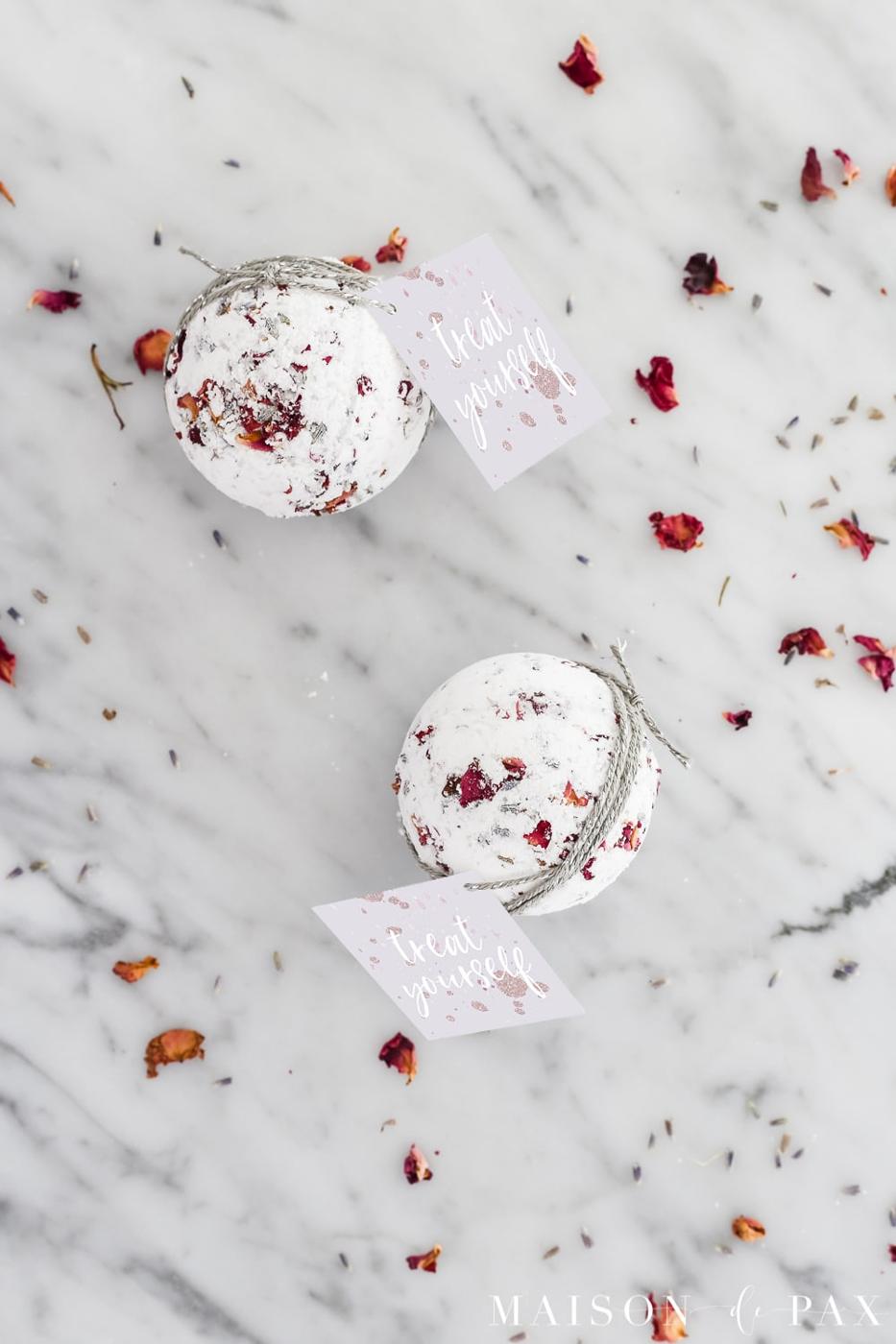 Two white bath bombs studded with dried rose petals and lavender, tied with silver cord and treat yourself tags on marble