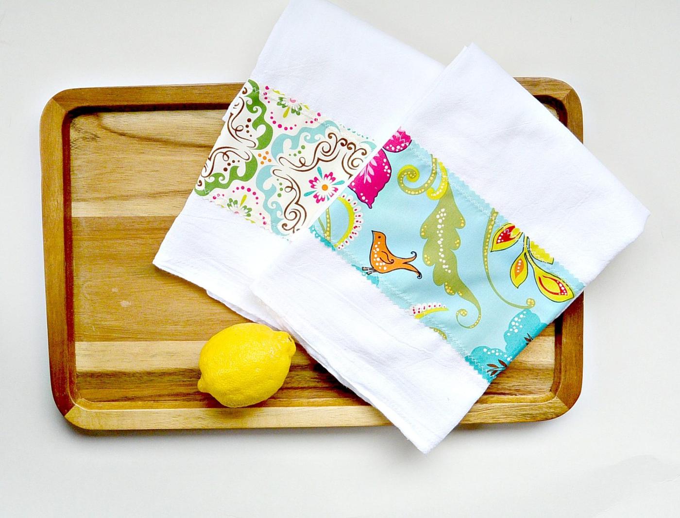 Two white flour sack tea towels with bands of colorful floral and bird print fabric stitched across them on a wood tray