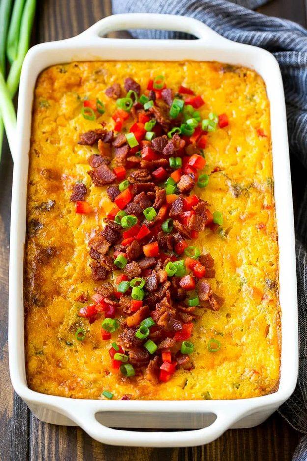 Corn casserole topped with bacon, cheddar, red peppers and green onions in a white handled baking dish
