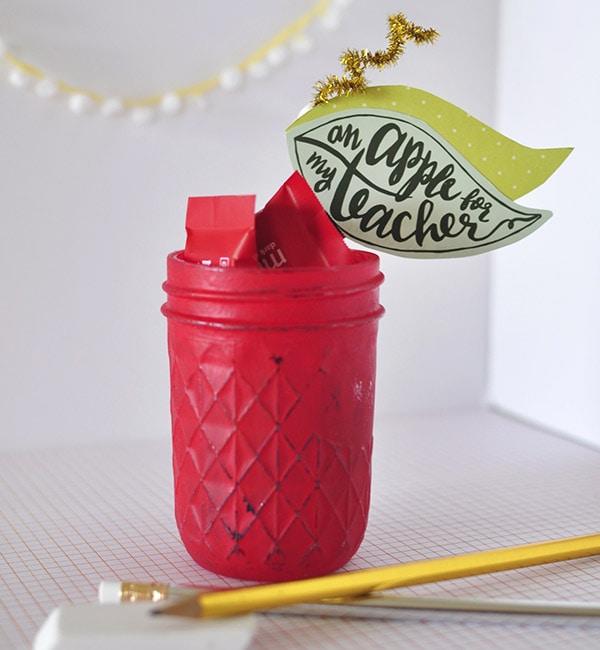Mason jar painted red as an apple with a green leaf and brown stem on the lid, filled with wrapped candy