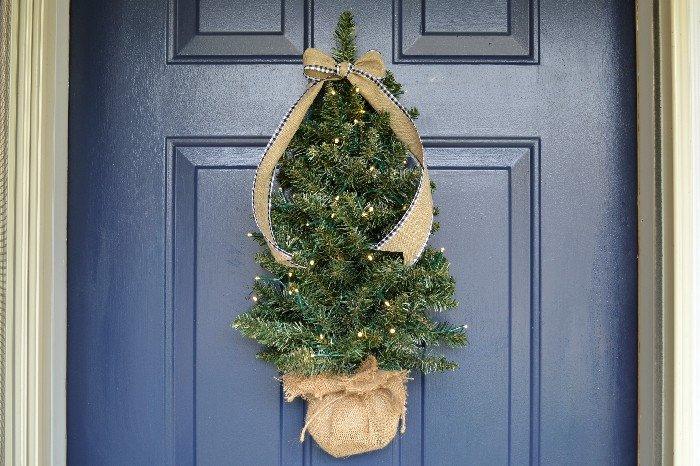 Small artificial Christmas tree hung flat against a front door with lights and ribbon woven through it