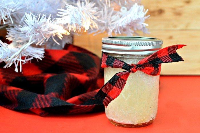 Small quilted glass jar of pale sugar scrub tied with red and black buffalo check ribbon beside a white tree