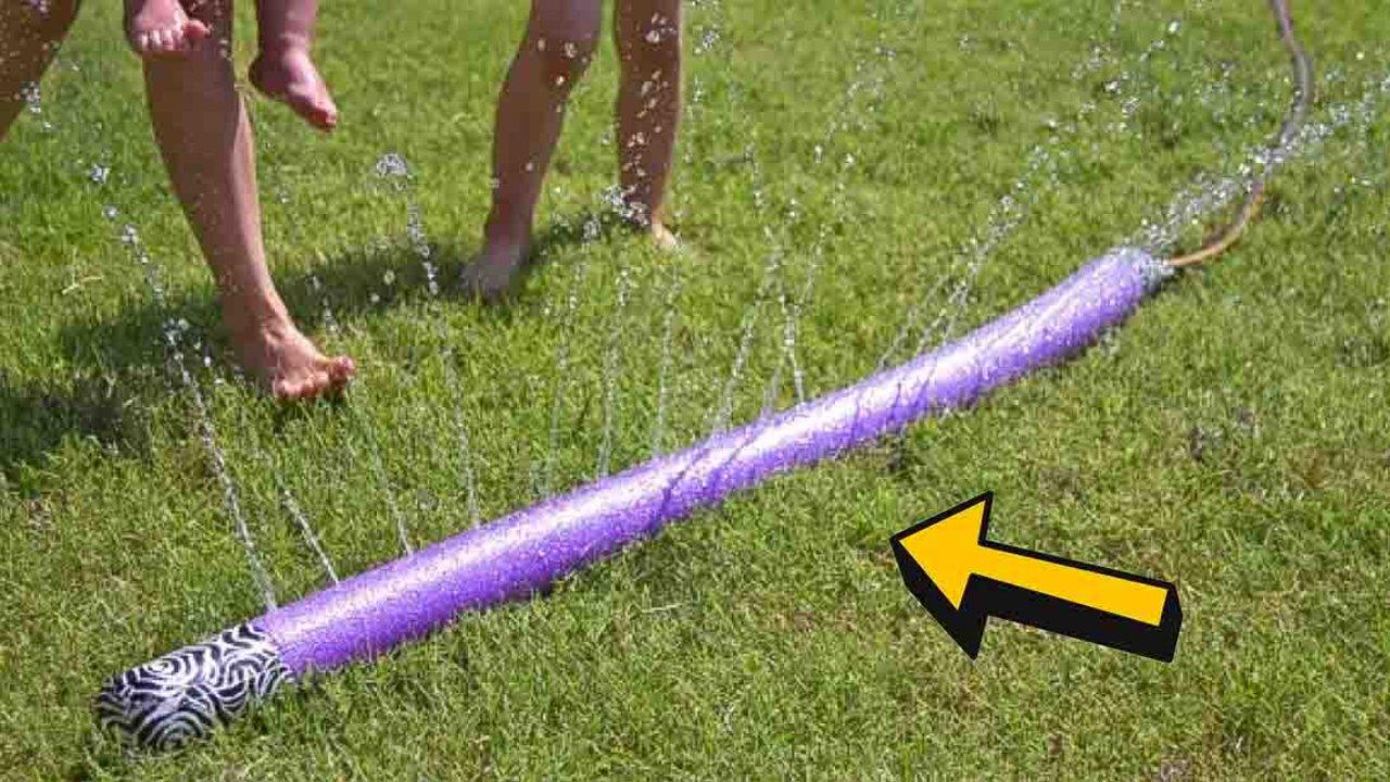 Purple pool noodle lying on grass with water jetting up from holes along its length while two children run through it
