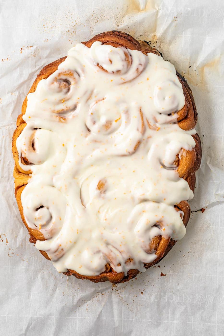 Orange sweet rolls baked together on parchment and covered with an orange-speckled glaze in a finished view
